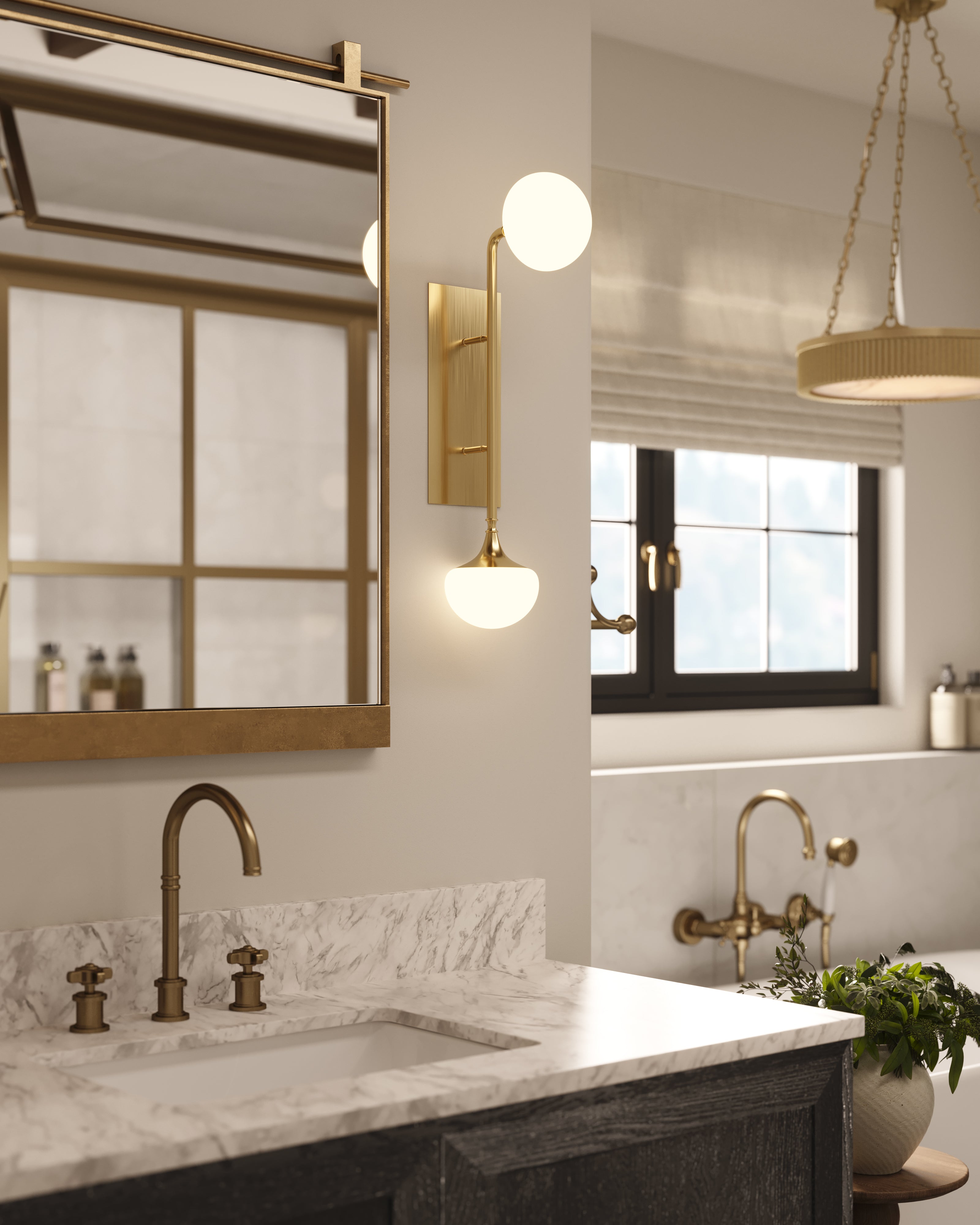Bathroom interior with a wall sconce in aged brass beside a mirror, marble countertop, brass fixtures, and a window with natural light.