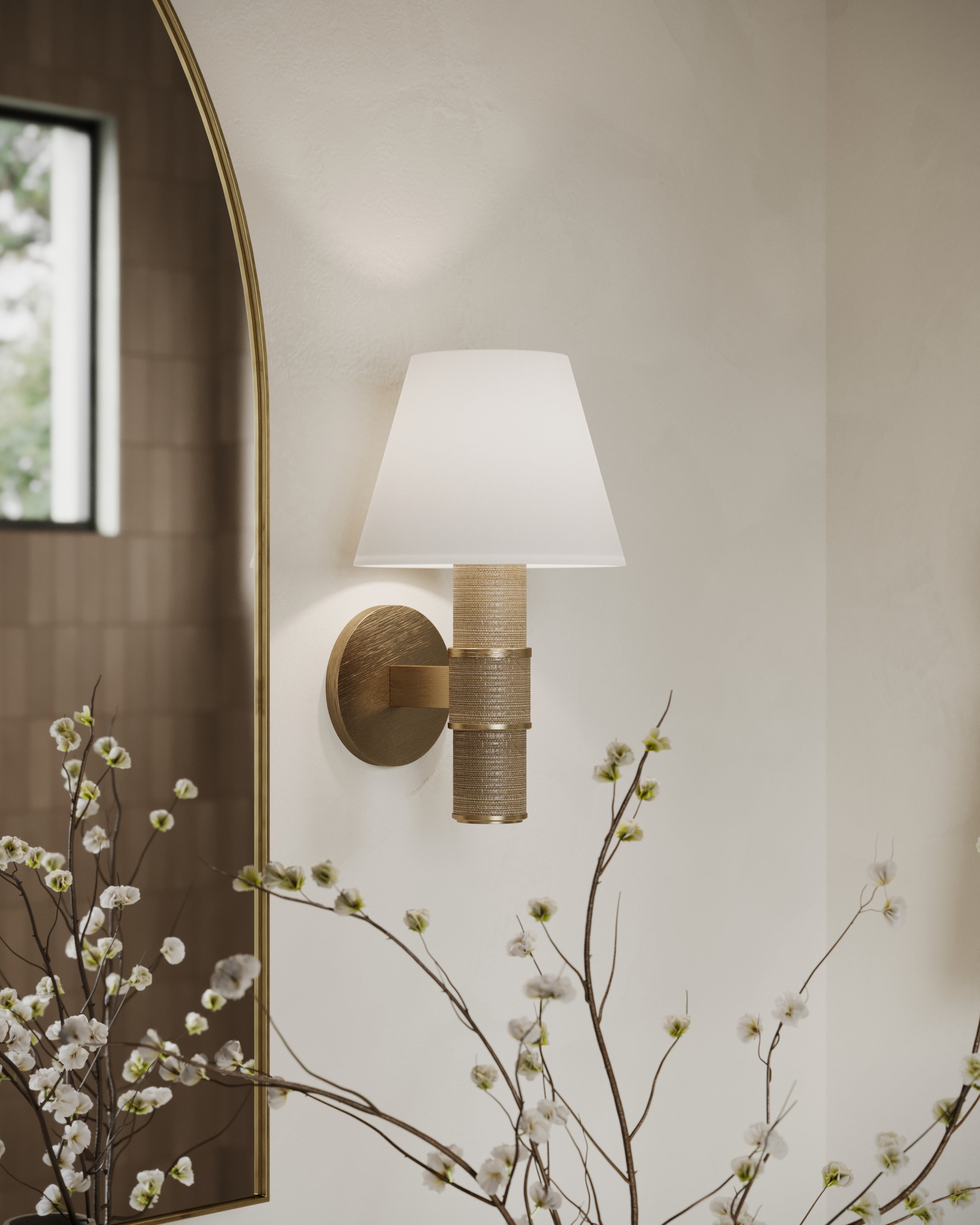 Wall-mounted patina brass sconce with woven grass cloth detail and white fabric shade, casting warm light beside a mirror.