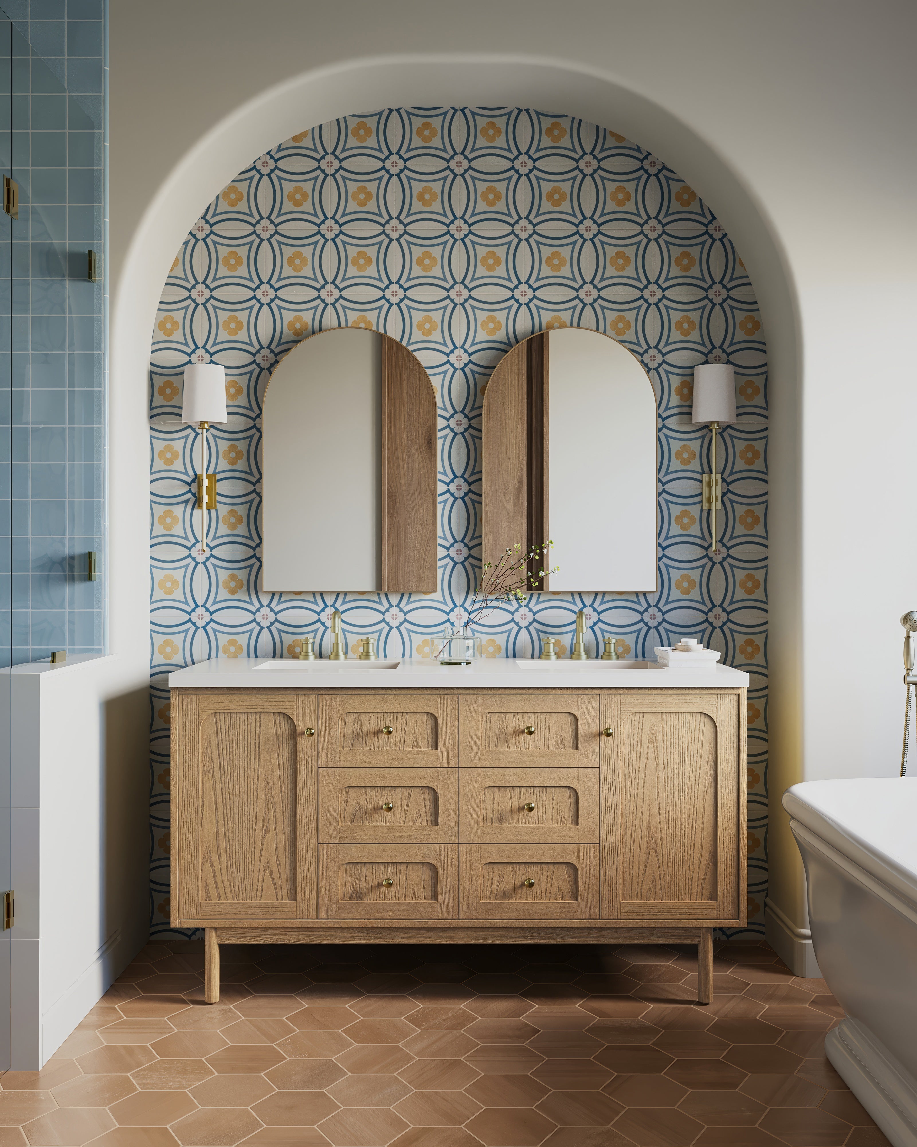 Double-sink vanity with arched brass mirrors and fixtures, and a floral tile accent wall set in an arched alcove with warm lighting.