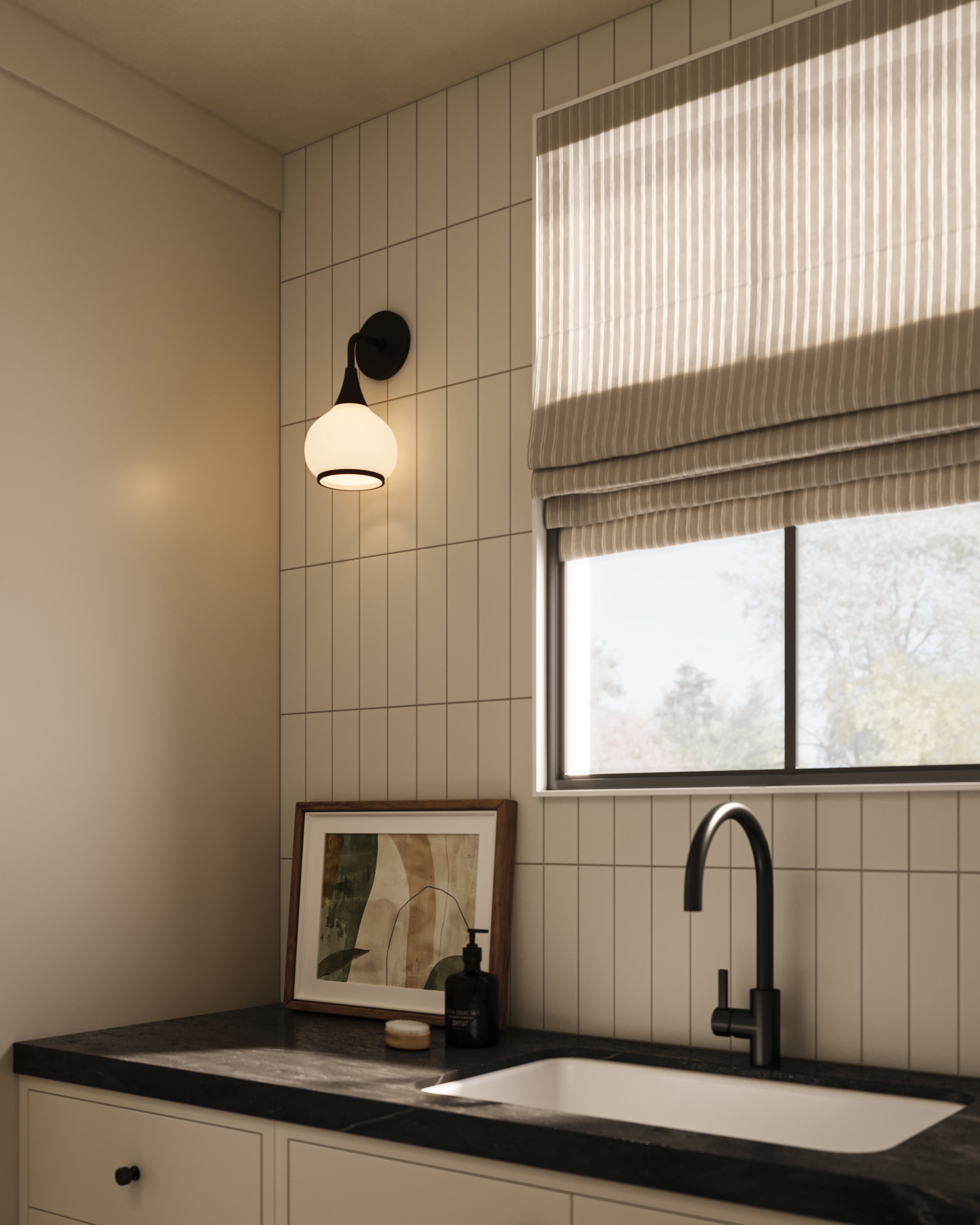 Matte black wall sconce with opal glass shade mounted on a vertically tiled wall above a bathroom sink and black countertop beside a window with striped Roman shade.
