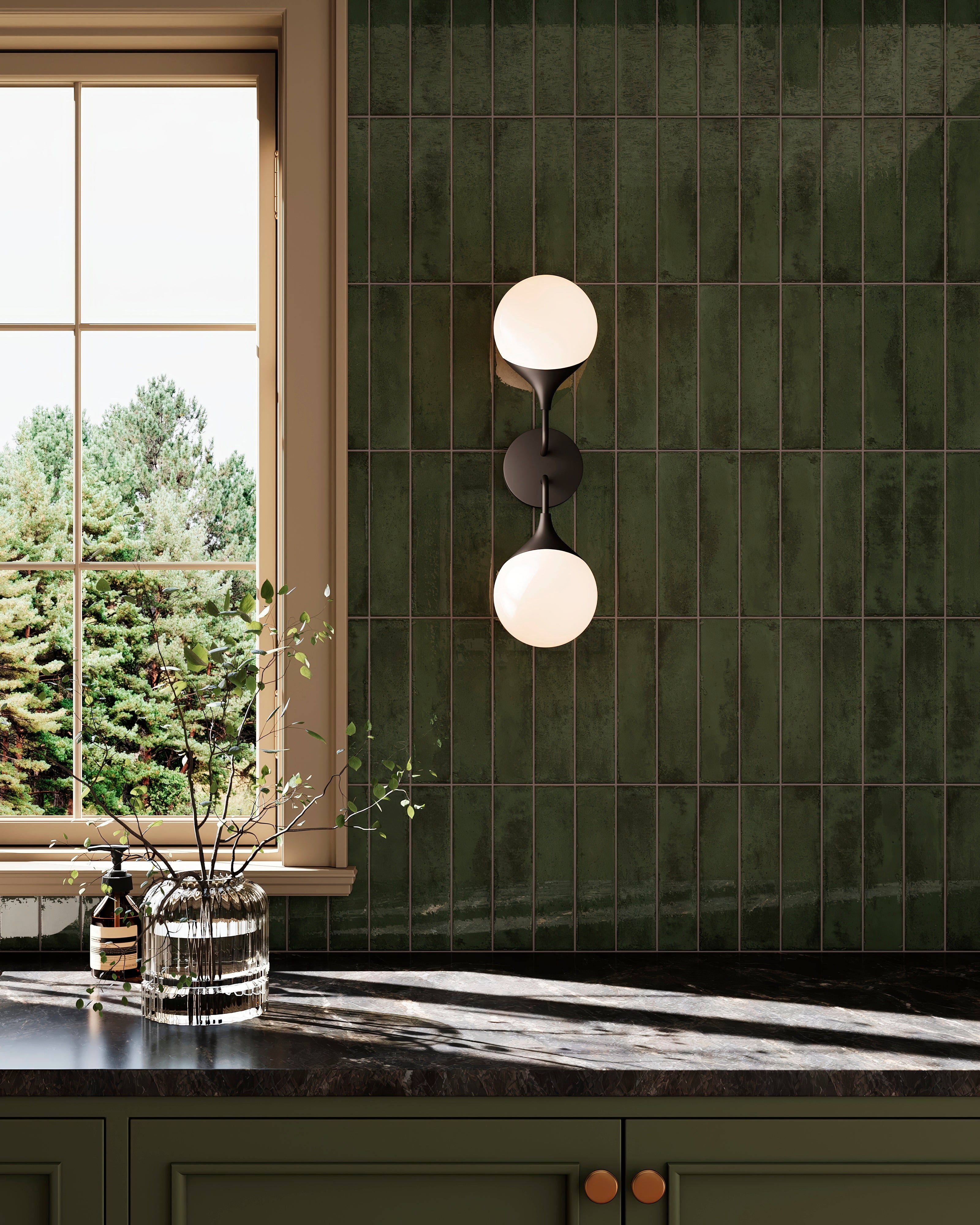 Matte black wall light with stacked frosted globes glowing against deep green tile walls beside a kitchen window and dark countertop with glass décor.
