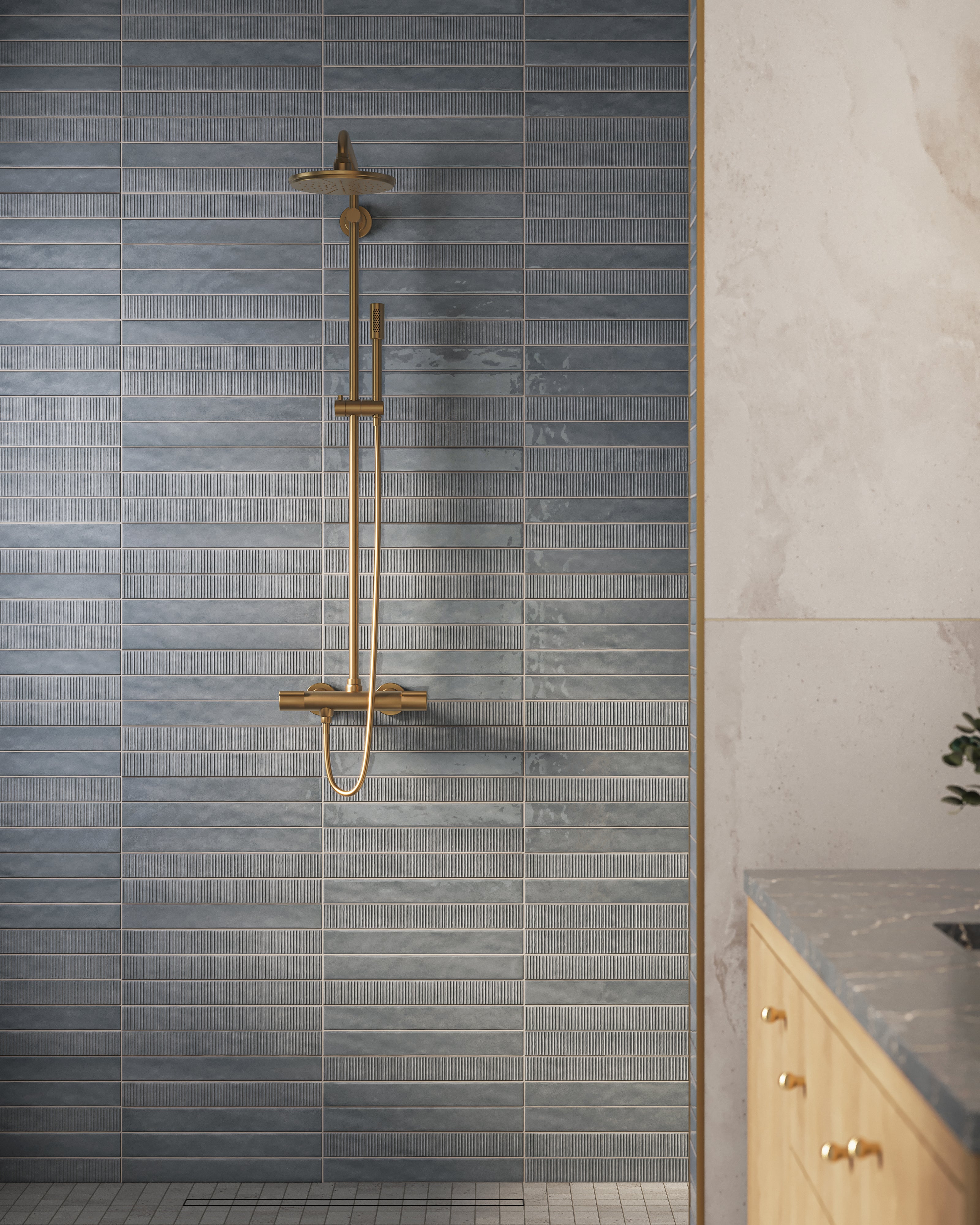 Close-up of a brass shower fixture mounted on gray-blue ribbed wall tiles beside a light wood vanity with a dark stone countertop.