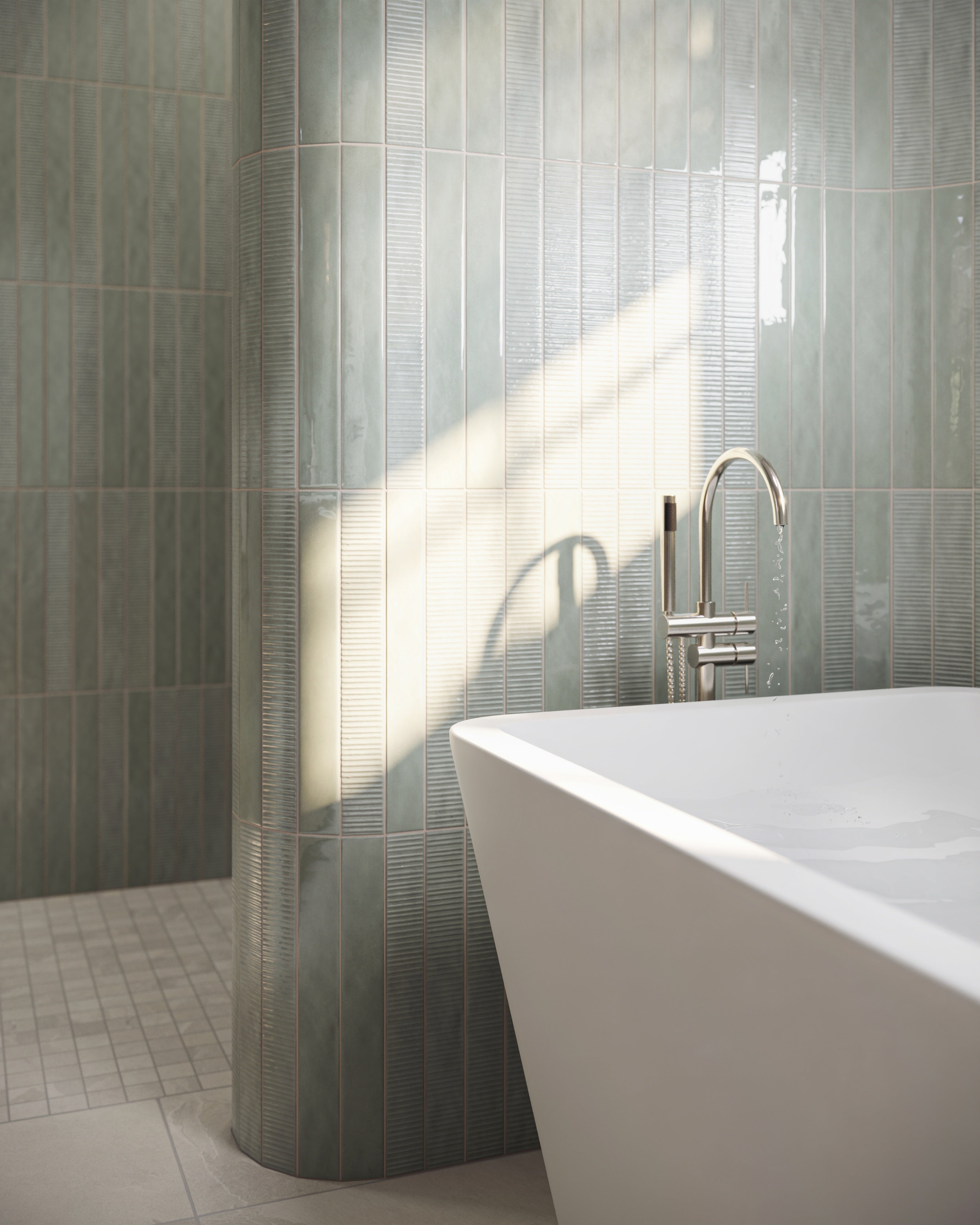 Freestanding white bathtub beside glossy pale green tiled walls with a curved tile column, chrome tub filler, and soft natural light.