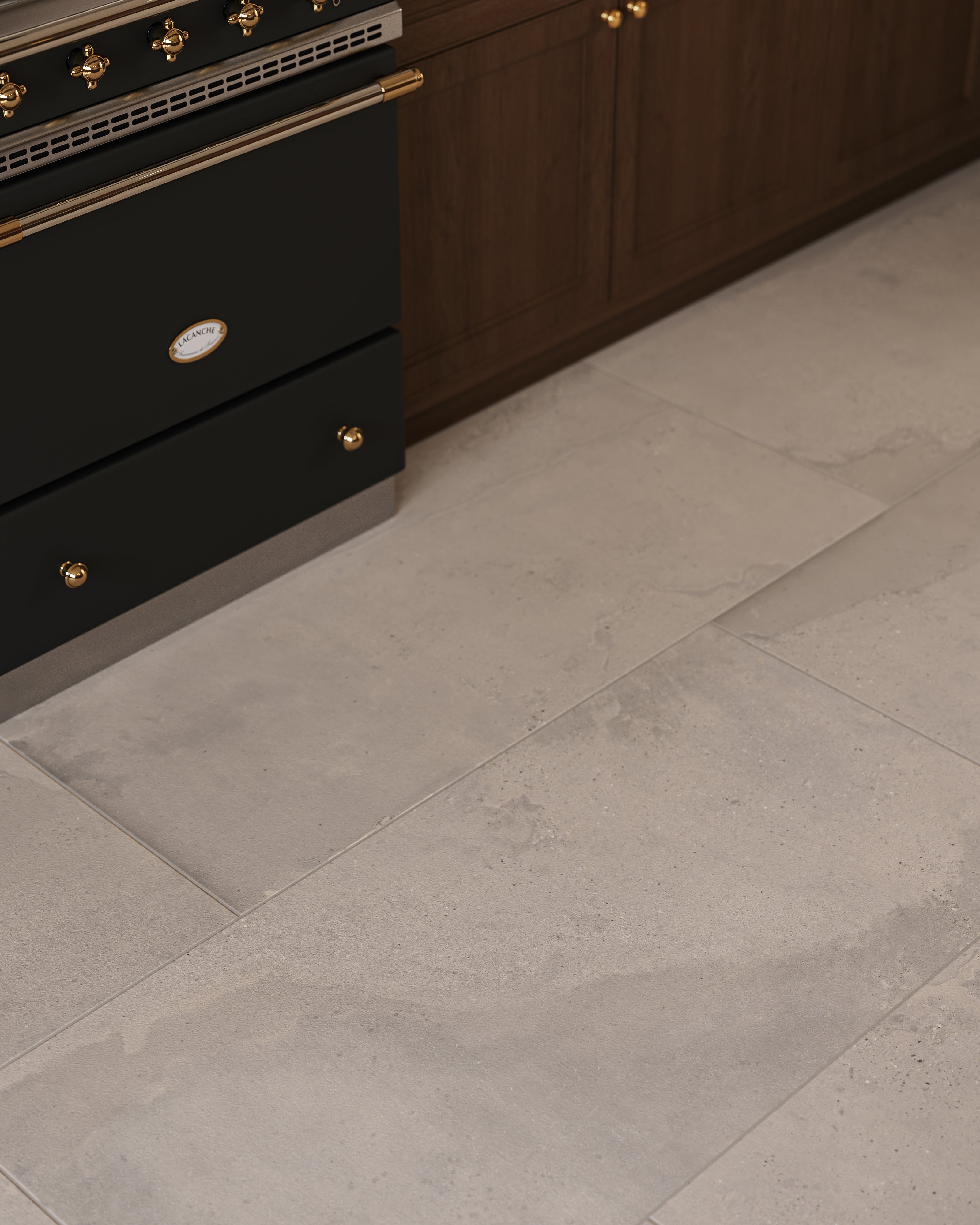 Kitchen floor close-up with taupe large-format rectangular tile showing soft mineral flecks and natural stone-like variation, set beside dark wood cabinetry and a black range with brass hardware.