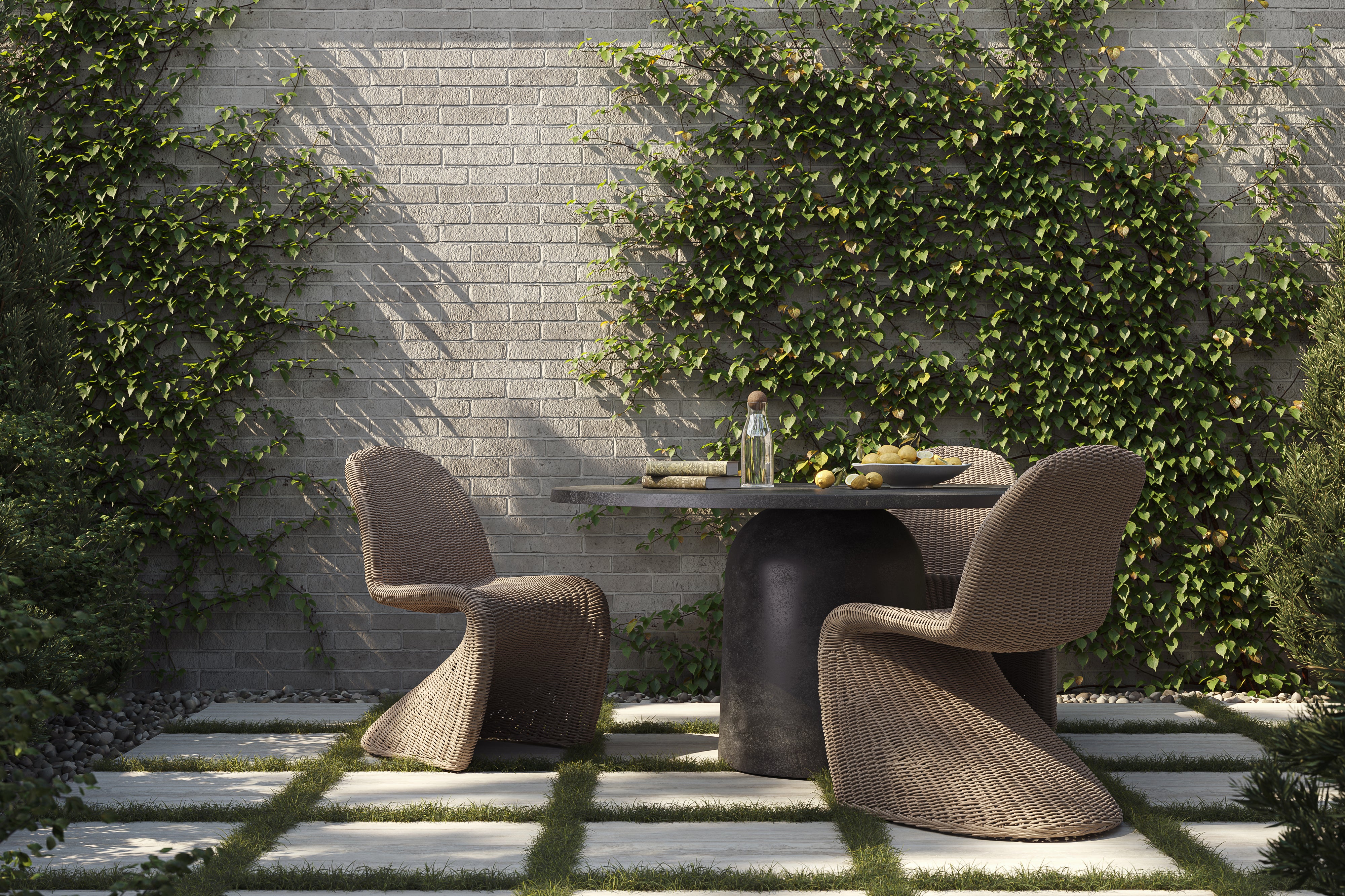 Outdoor garden dining area with a round aged dark gray pedestal table set on square pavers with grass joints, paired with two woven chairs against an ivy-covered brick wall and surrounding greenery.