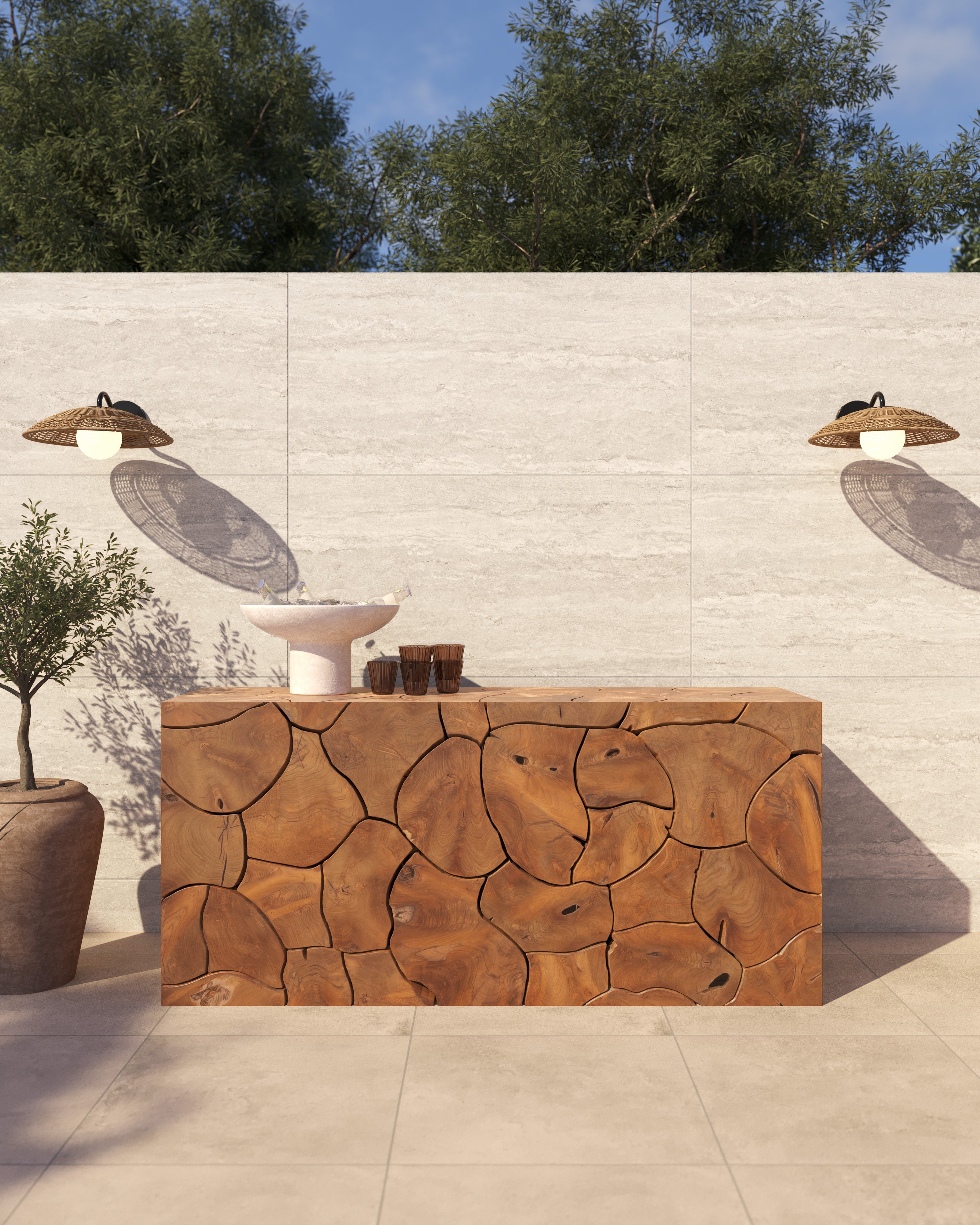 Our Cielo Outdoor Console Table topped with white ceramic vessels and dark wood accents, set against a cream stone wall with two wicker sconces and a potted olive tree beside it.