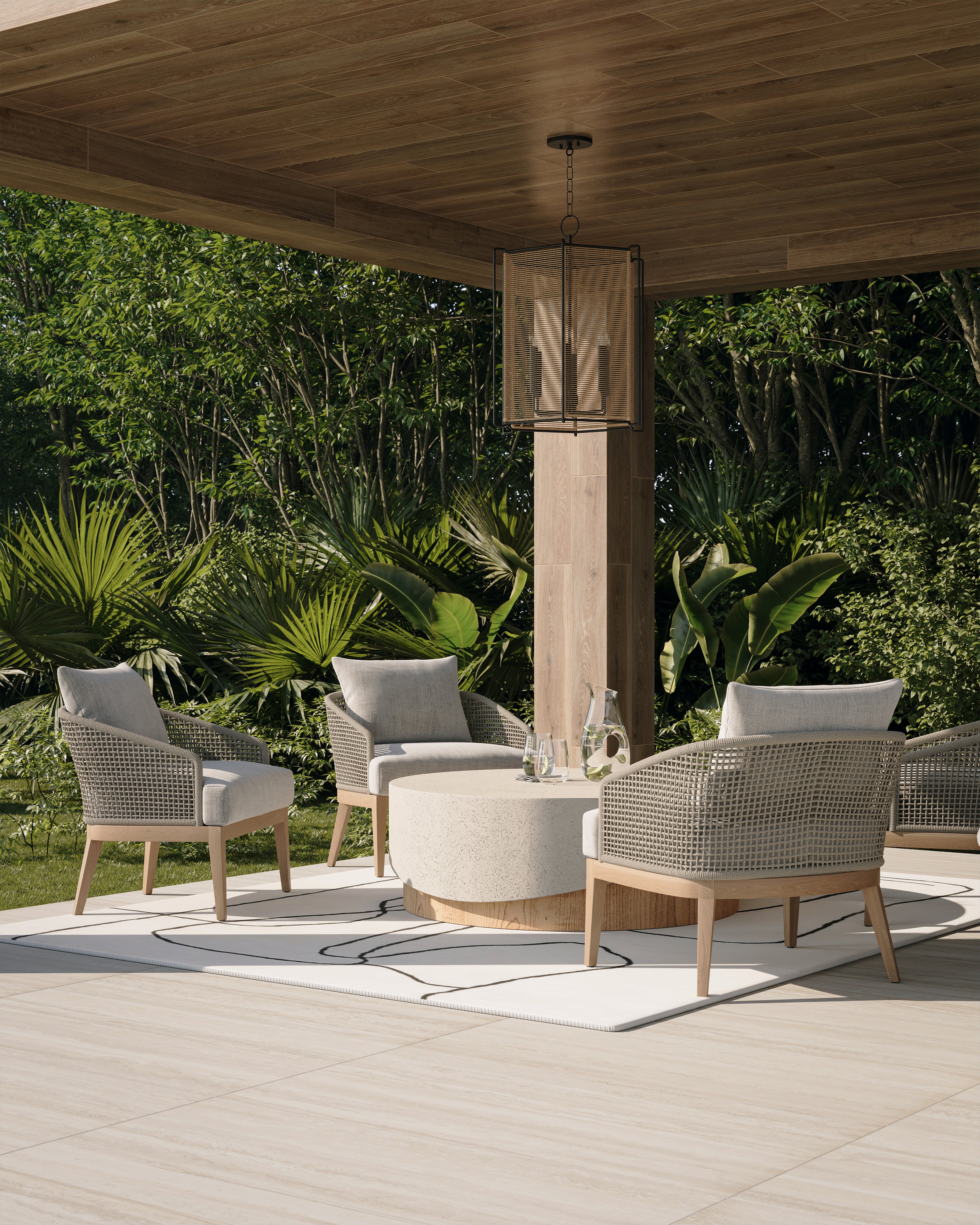 Covered outdoor lounge with cream cushioned rope chairs, light wood frames, a white abstract-line rug, round stone coffee table, wood-look ceiling tile, pale floor tile, tropical greenery, and a black-framed hanging lantern.