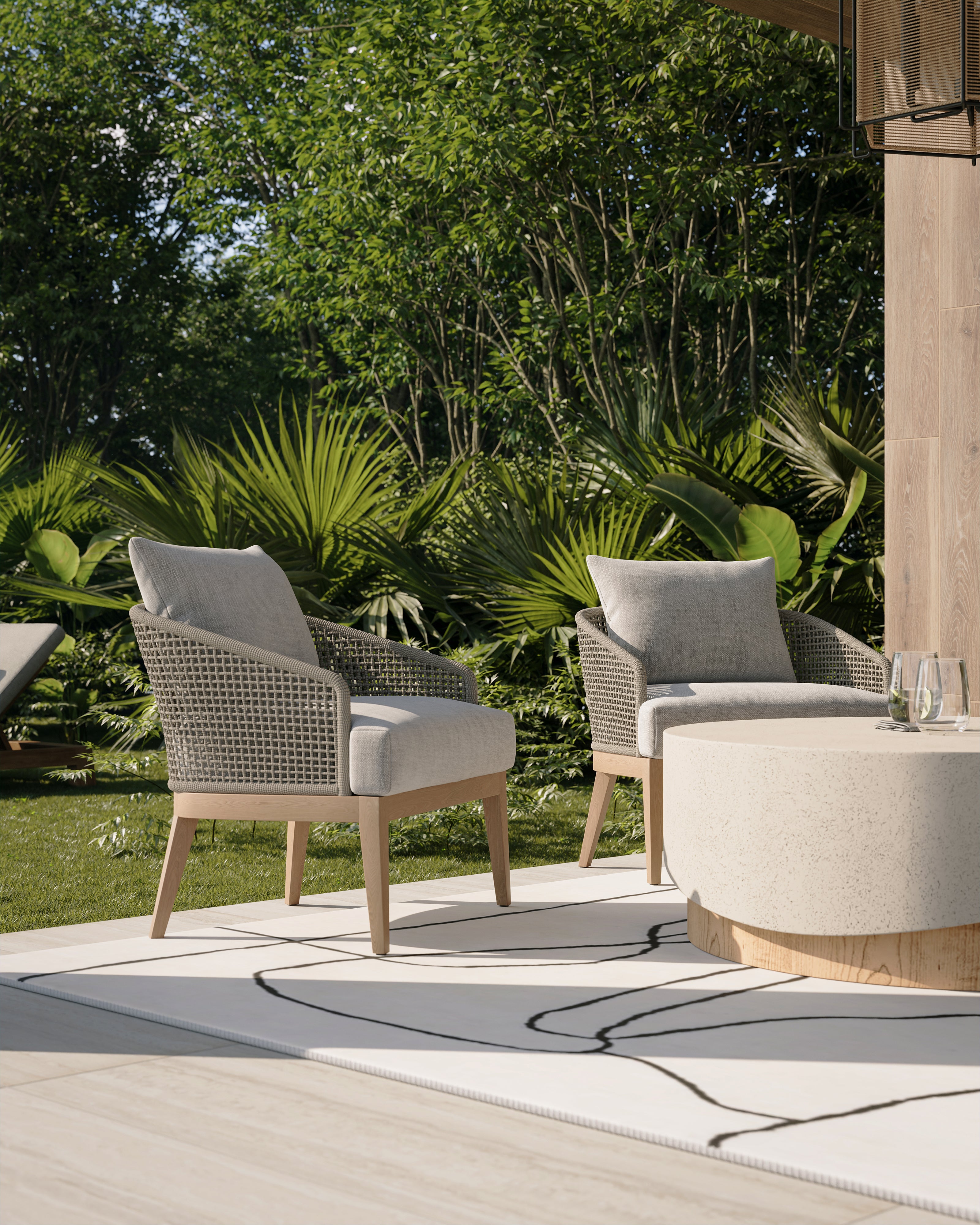 Concrete round outdoor coffee table with a textured beige top and warm wood look base, paired with woven outdoor lounge chairs in a lush garden setting.