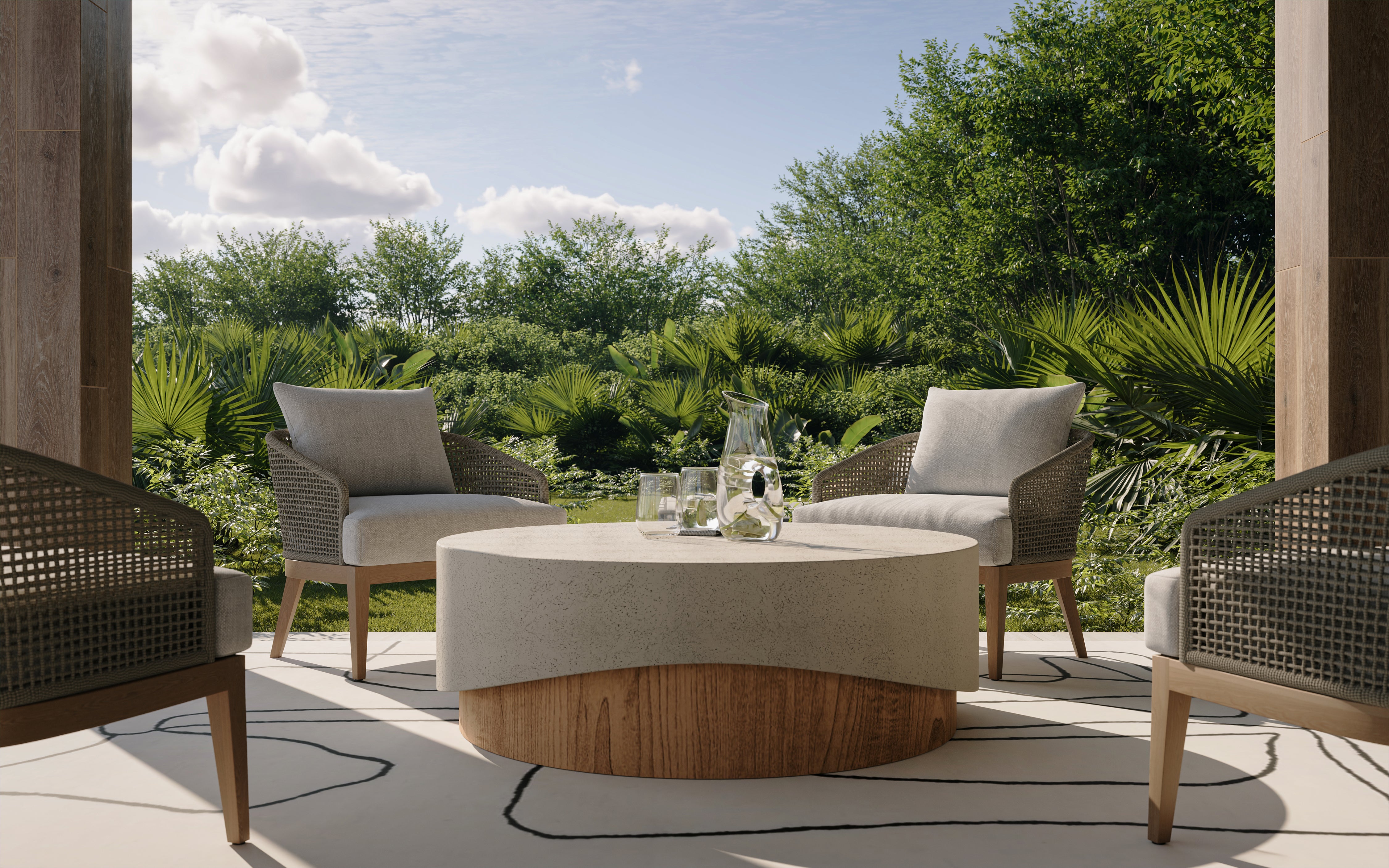 Concrete round outdoor coffee table with a textured beige top and warm wood look base, styled on an open-air patio with woven lounge chairs and lush greenery.