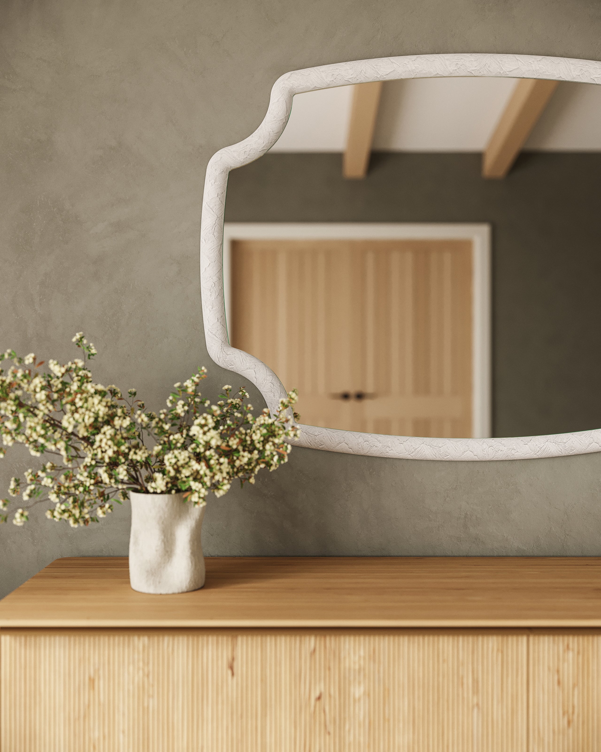 White textured Seraphin mirror with sculpted resin frame displayed above a wood dresser, reflecting warm ceiling beams in a serene interior.
