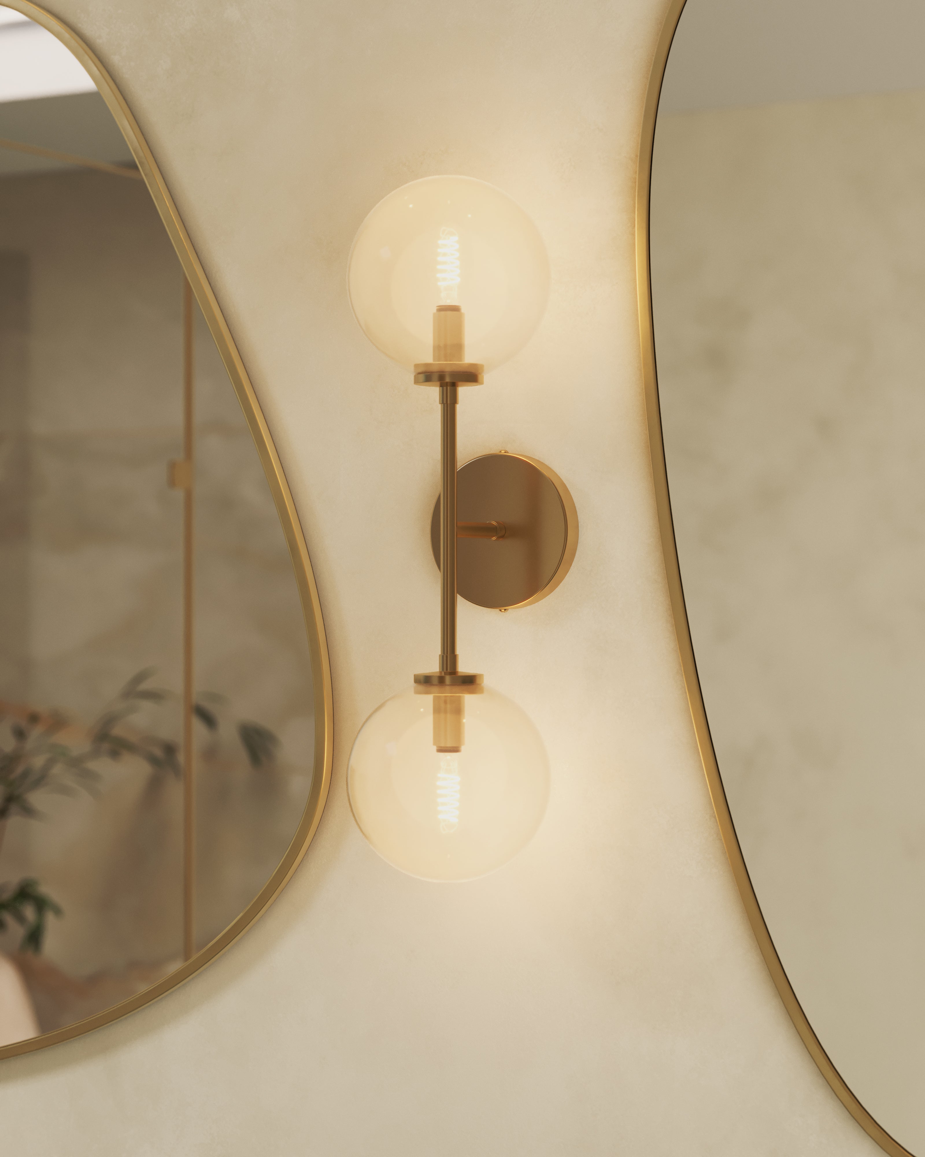 Modern bathroom wall sconce with dual clear glass globe shades and brushed brass arm beside an asymmetrical mirror.
