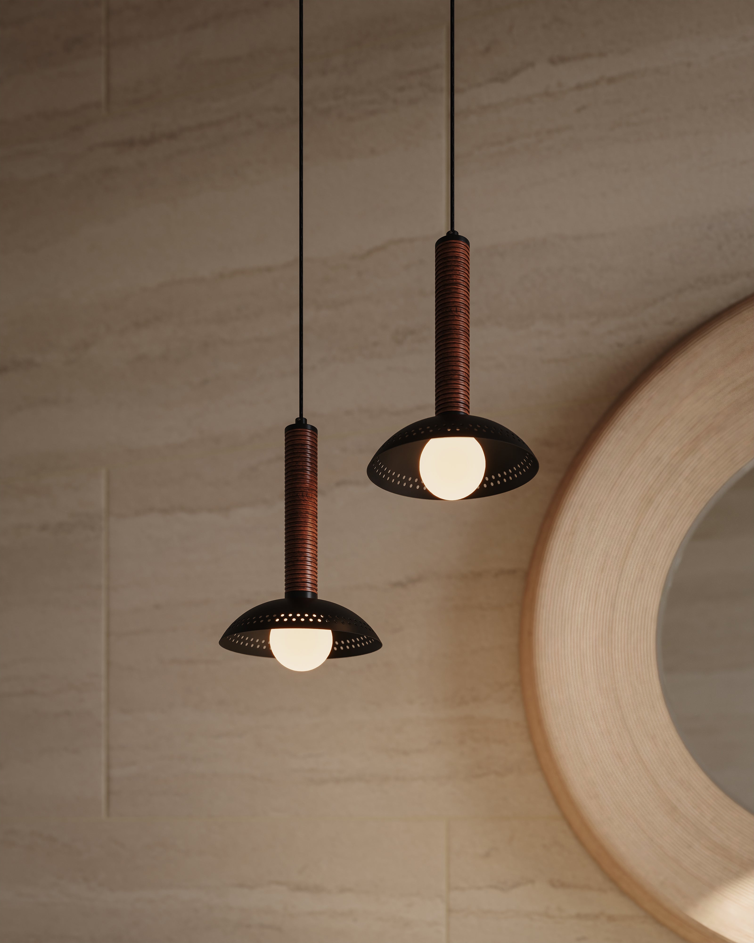 Pair of black pendants with ribbed brown stems and shallow dome shades, softly glowing against a light stone wall beside a round mirror