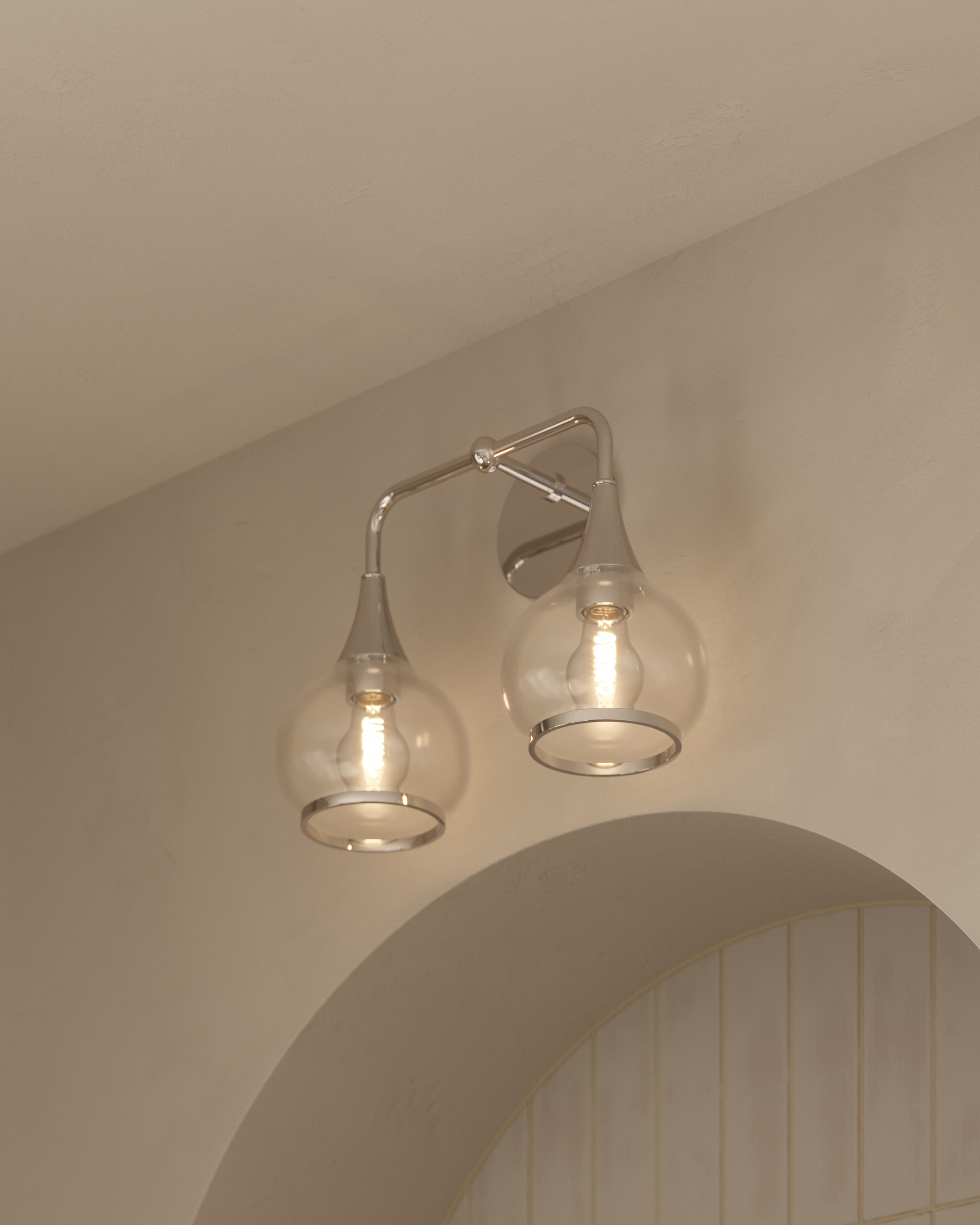 Chrome double wall sconce with curved arm and clear glass globe shades, illuminated above an arched mirror.