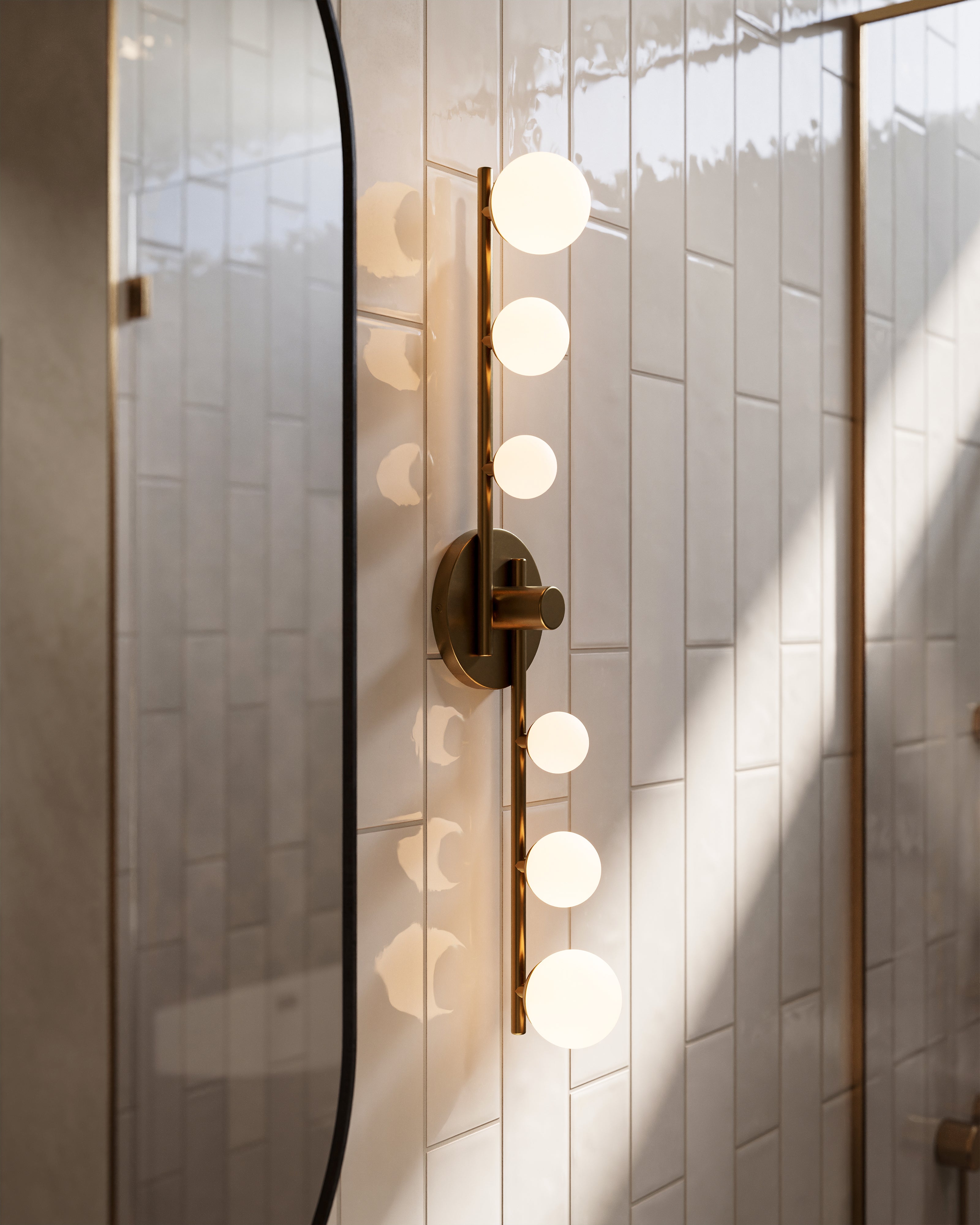 Brushed gold LED vanity light with frosted globe bulbs mounted beside an arched bathroom mirror, casting warm light across glossy white vertical tiles and soft shadows.