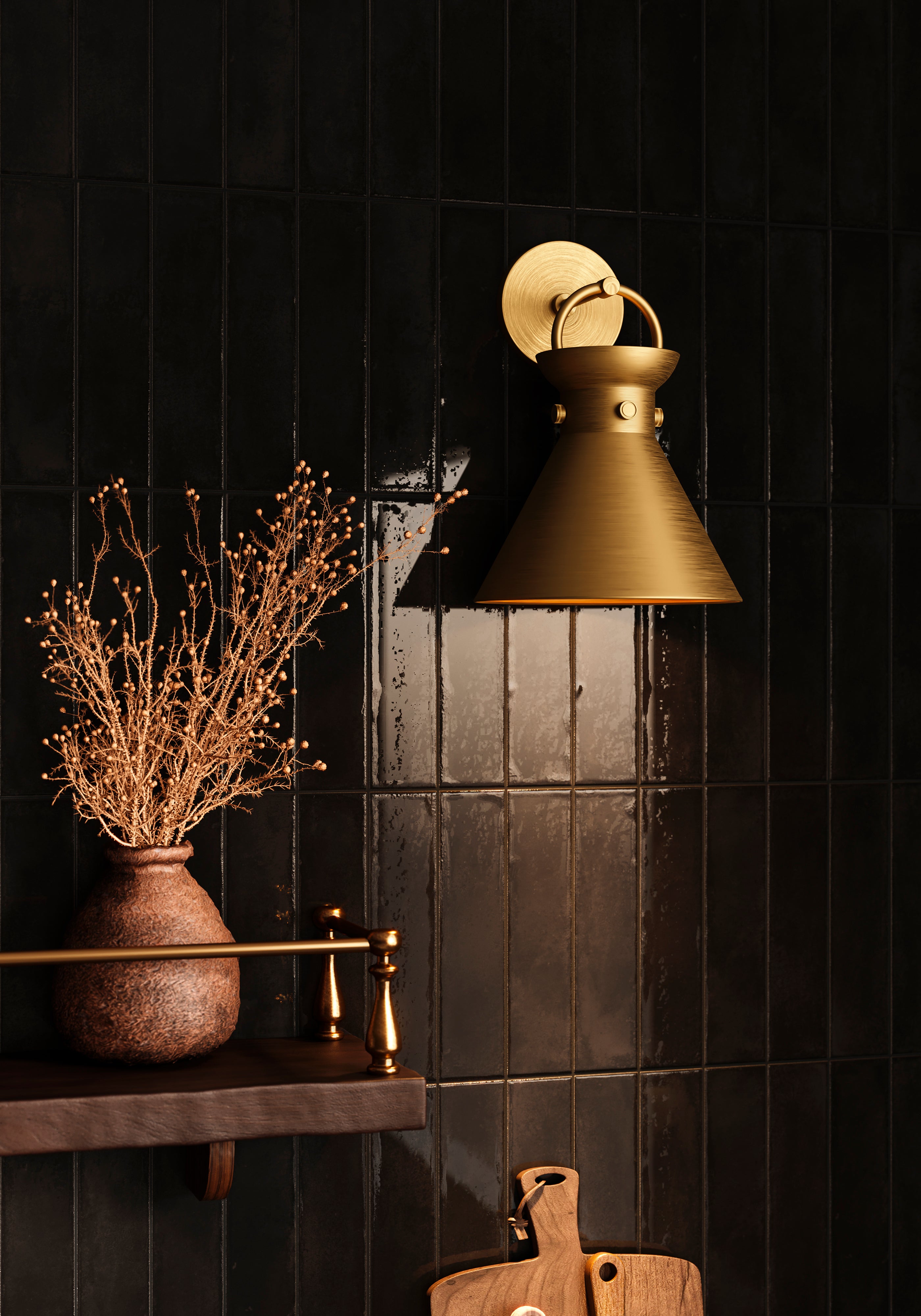 Aged gold wall sconce with conical metal shade mounted on a black tiled wall above a wooden shelf with a ceramic vase and dried branches.