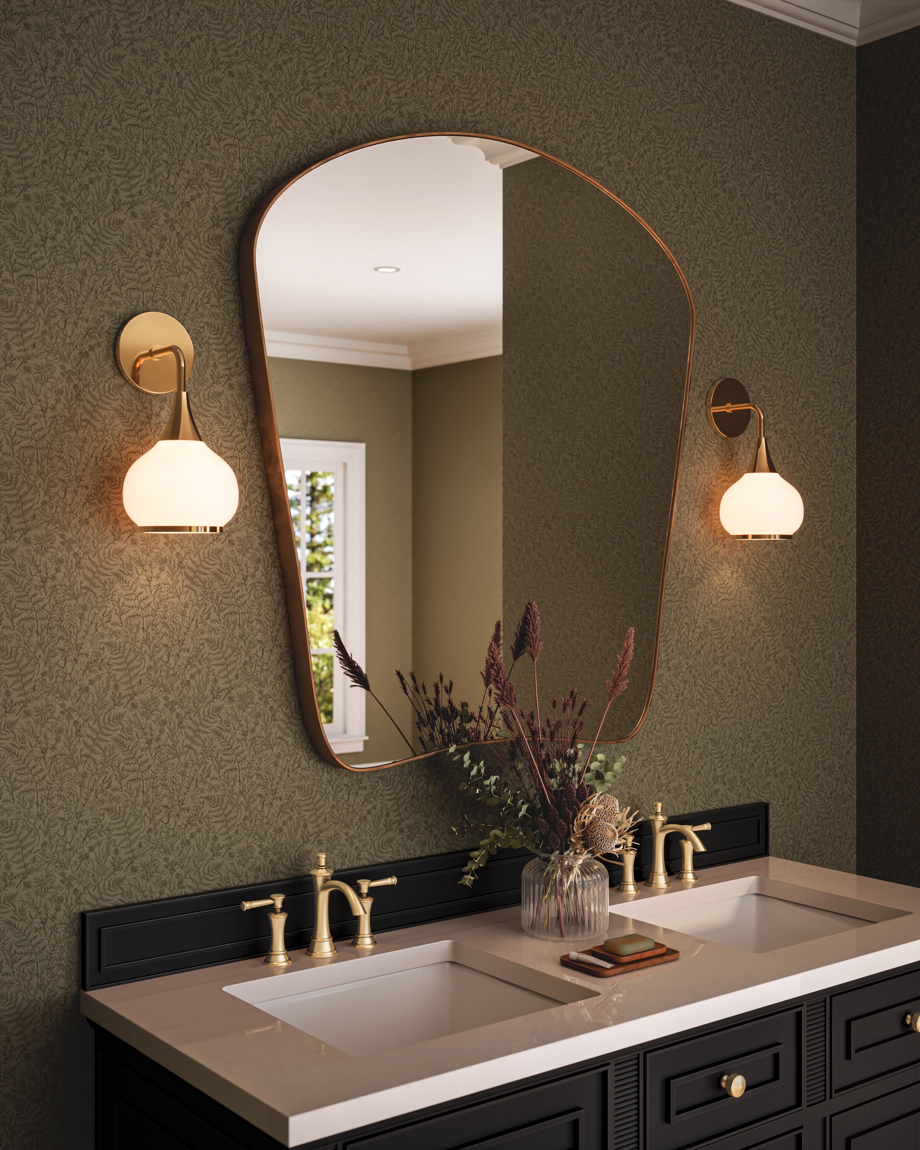 Bathroom vanity with olive green botanical wallpaper, featuring a black double vanity with brass faucets, white sinks, a sculptural asymmetrical mirror, and warm brass wall sconces illuminating decorative dried florals.