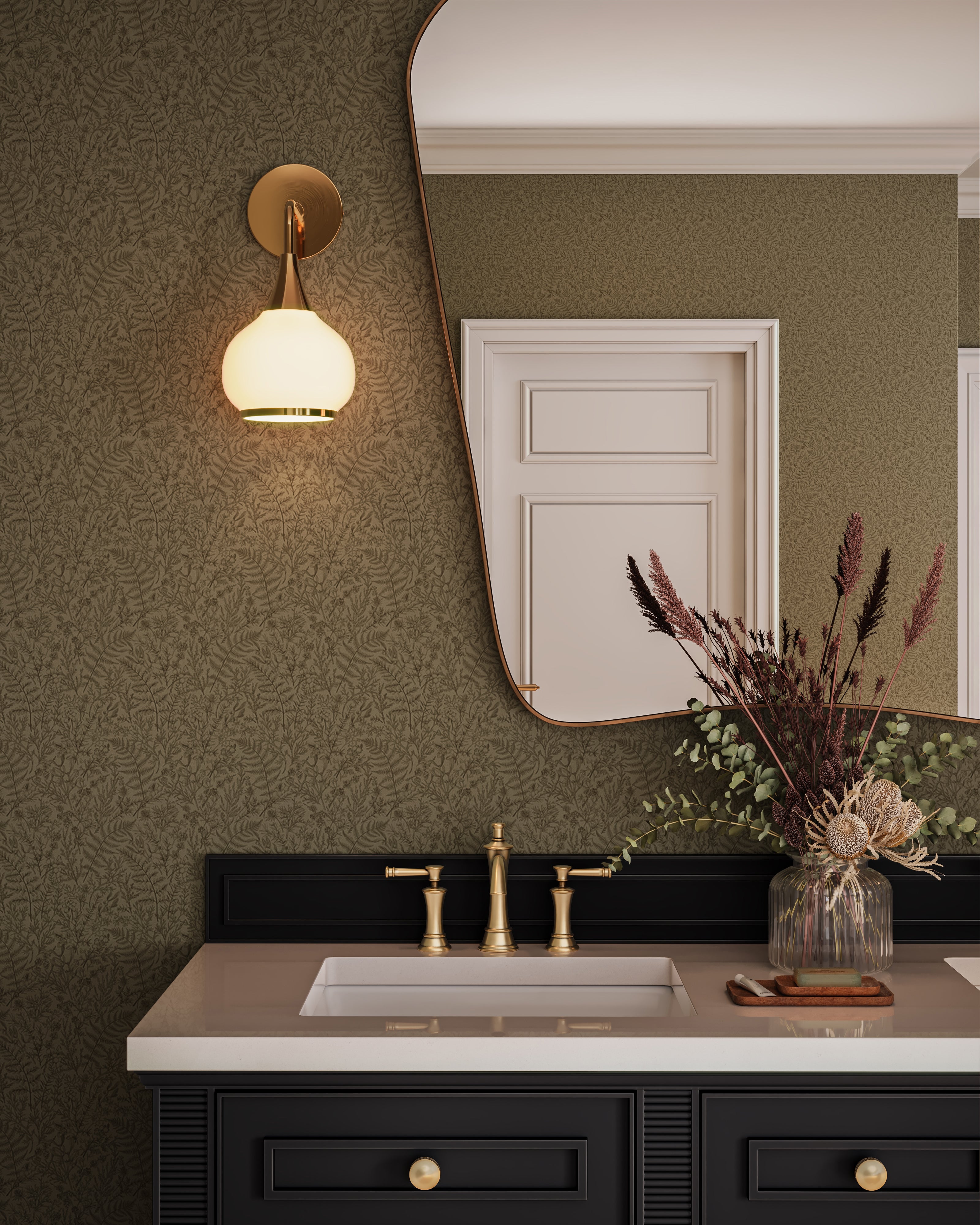 Elegant bathroom vanity styled with olive green botanical wallpaper, a black vanity cabinet with brass hardware, a white countertop sink with brass faucet, a warm wall sconce, and an asymmetrical mirror reflecting a paneled door and decorative dried florals.
